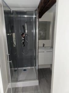 Salle de bain moderne avec cabine de douche vitrée et panneau de contrôle multipositions, face à un meuble vasque blanc sous un miroir encore partiellement protégé par un film plastique.