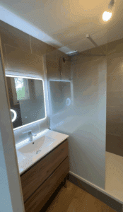 Salle de bain moderne avec meuble vasque en bois, lavabo blanc, robinet chromé et miroir lumineux LED. Devant, une paroi de douche vitrée.